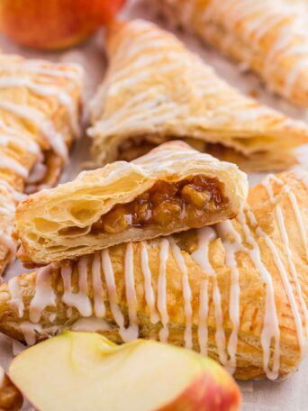 Flaky apple turnovers are drizzled with white icing. One turnover is cut open to reveal a spiced apple filling. A fresh apple is partially visible in the background.