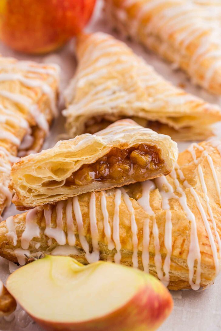 Flaky apple turnovers are drizzled with white icing. One turnover is cut open to reveal a spiced apple filling. A fresh apple is partially visible in the background.