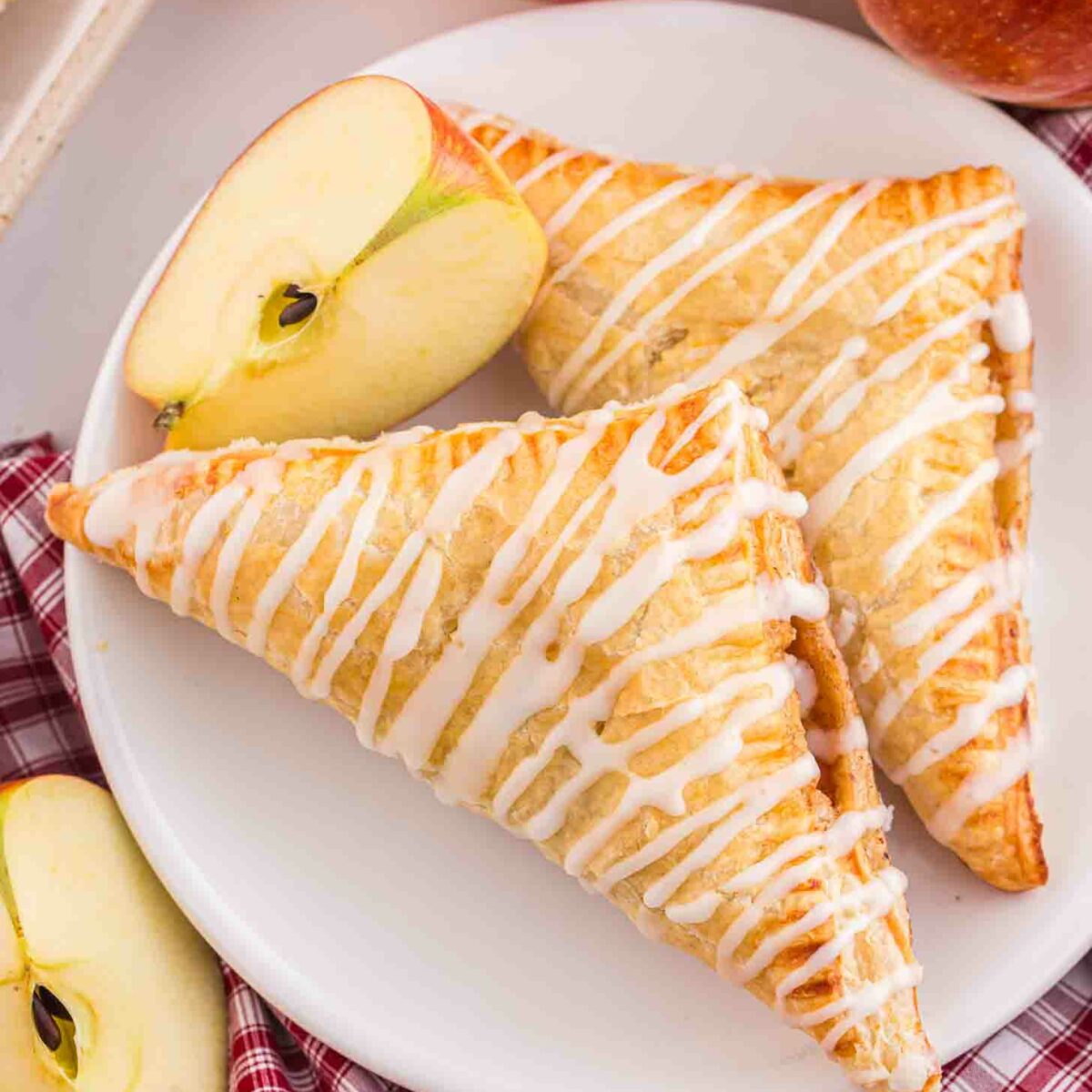 Two glazed apple turnovers on a white plate, topped with icing drizzle. The plate is on a red plaid cloth with sliced apples around it. Whole red apples are in the background.