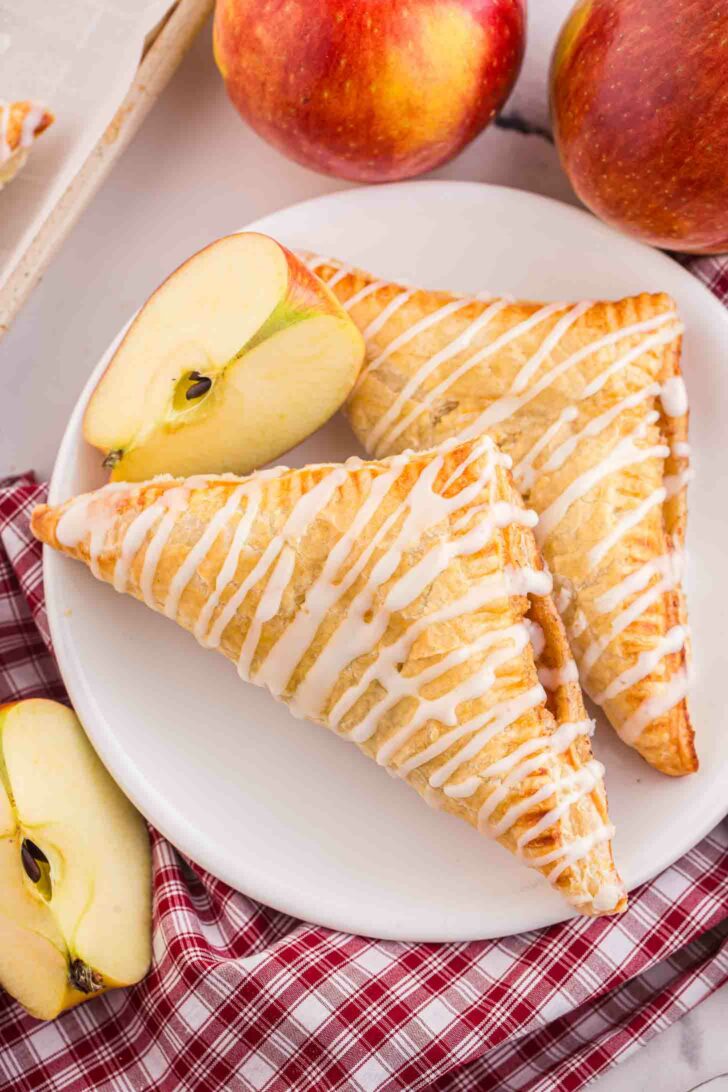 Two glazed apple turnovers on a white plate, topped with icing drizzle. The plate is on a red plaid cloth with sliced apples around it. Whole red apples are in the background.