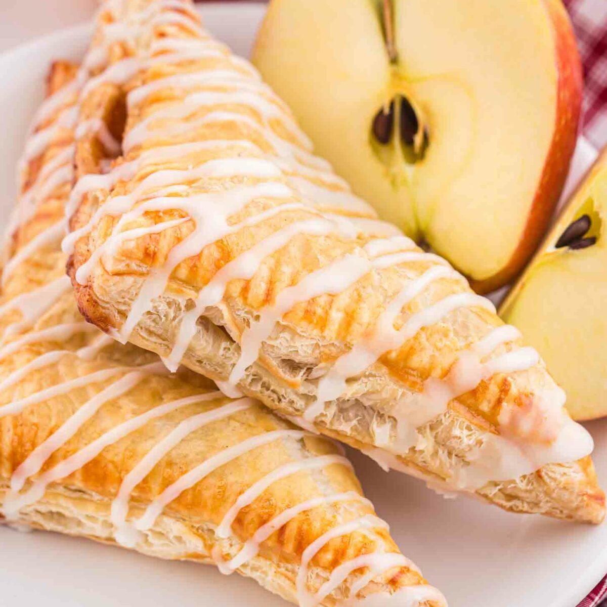 Two golden-brown apple turnovers drizzled with white icing are stacked on a white plate. Behind them, two red apples and an apple half are placed on a plaid cloth, creating a cozy, inviting scene.