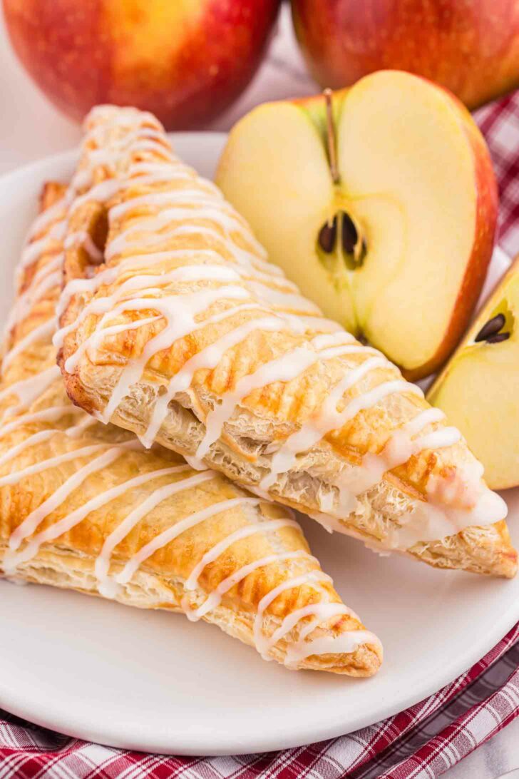 Two golden-brown apple turnovers drizzled with white icing are stacked on a white plate. Behind them, two red apples and an apple half are placed on a plaid cloth, creating a cozy, inviting scene.