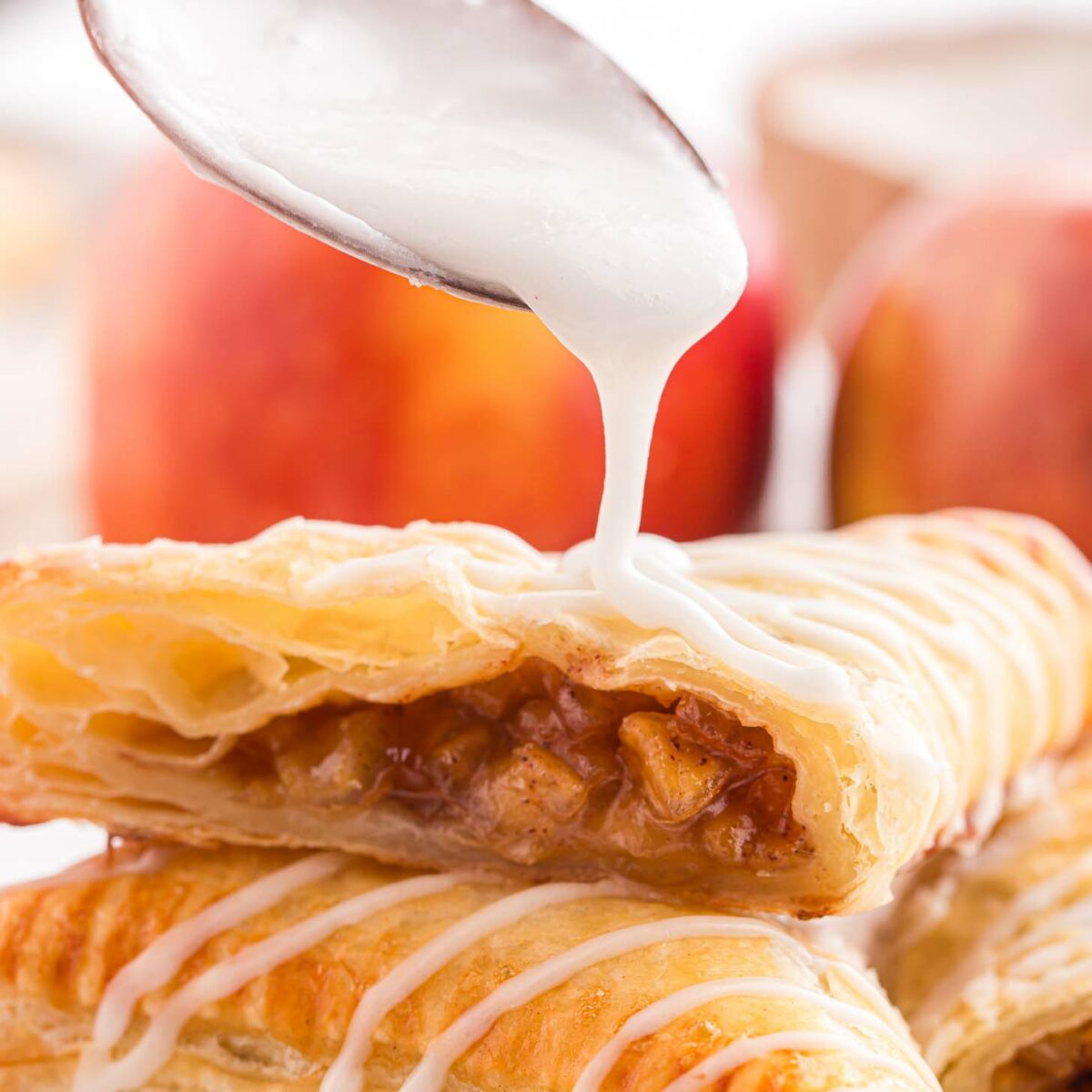 A spoon drizzles a powdered sugar glaze over flaky apple turnovers, with visible apple filling. Fresh apples are blurred in the background, enhancing the warm, inviting dessert scene.