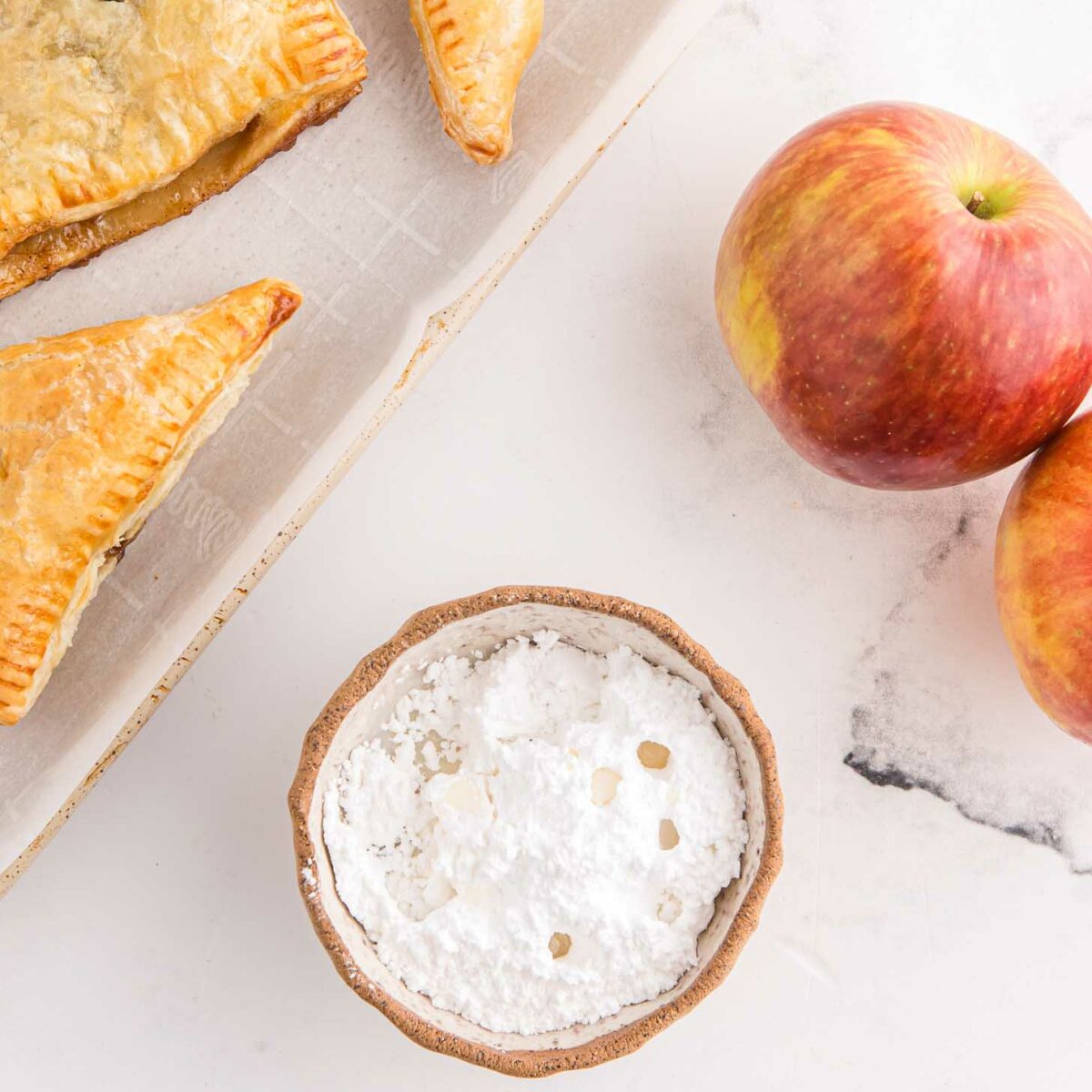 Freshly baked triangular pastries on a parchment-lined tray are next to a small bowl of powdered sugar and two red apples on a white marble surface.