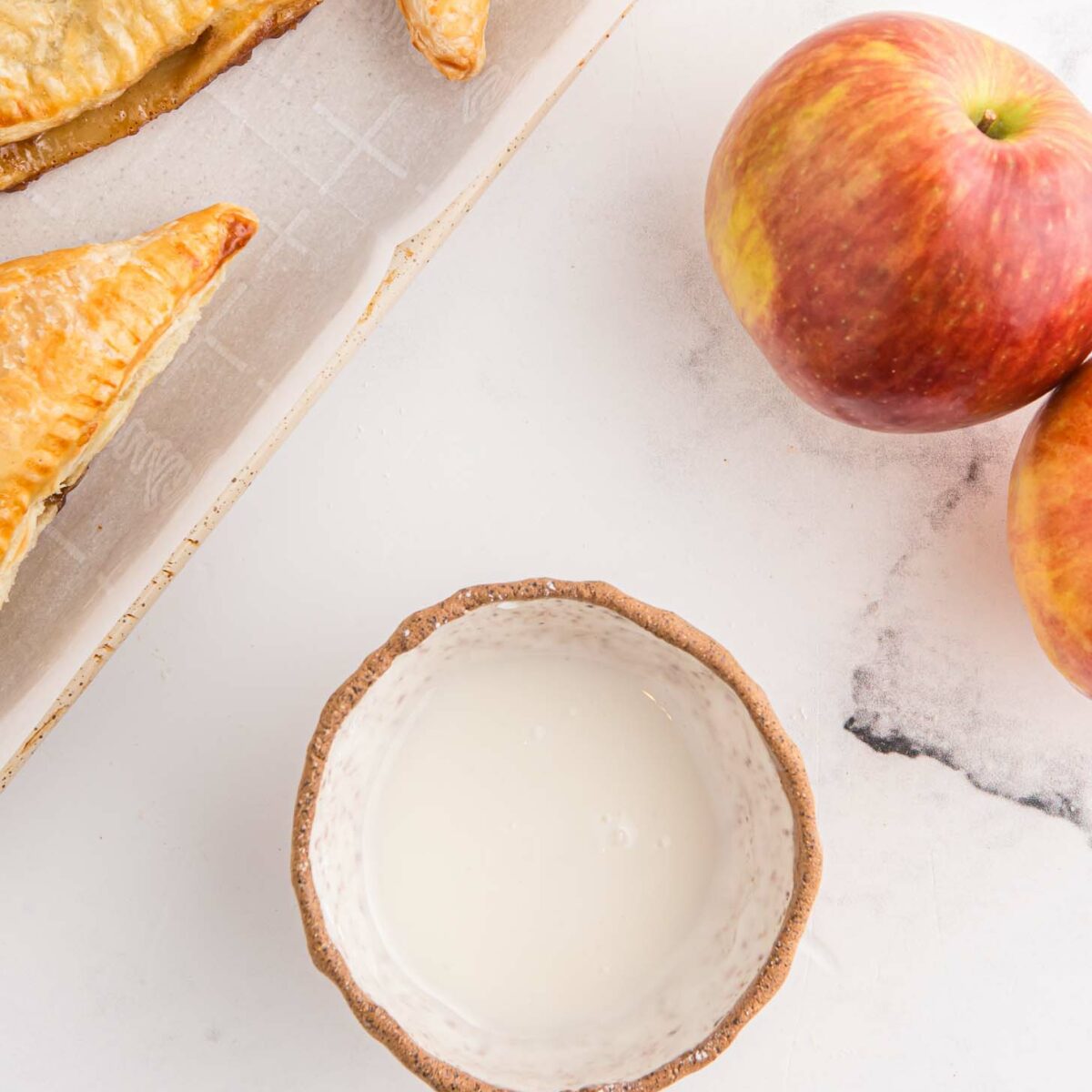 A wooden bowl filled with a powdered sugar glaze sits on a marble surface next to two red apples. Flaky golden-brown turnovers rest on a parchment-lined baking sheet to the left.