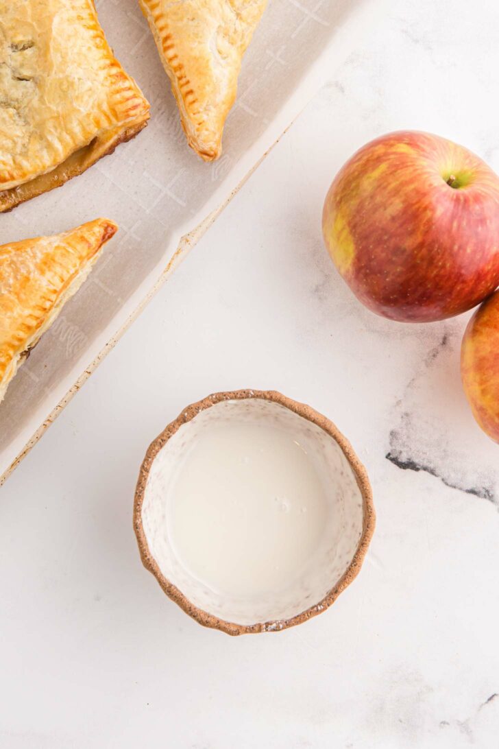 A wooden bowl filled with a powdered sugar glaze sits on a marble surface next to two red apples. Flaky golden-brown turnovers rest on a parchment-lined baking sheet to the left.