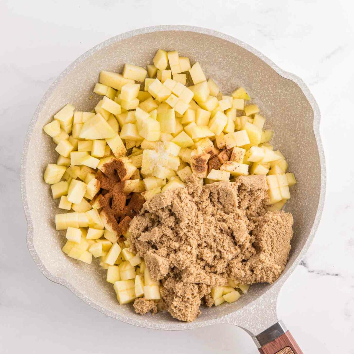 A frying pan filled with diced apples, brown sugar, and a sprinkle of cinnamon, all ready to be cooked. The pan is on a marble countertop with a wooden handle.