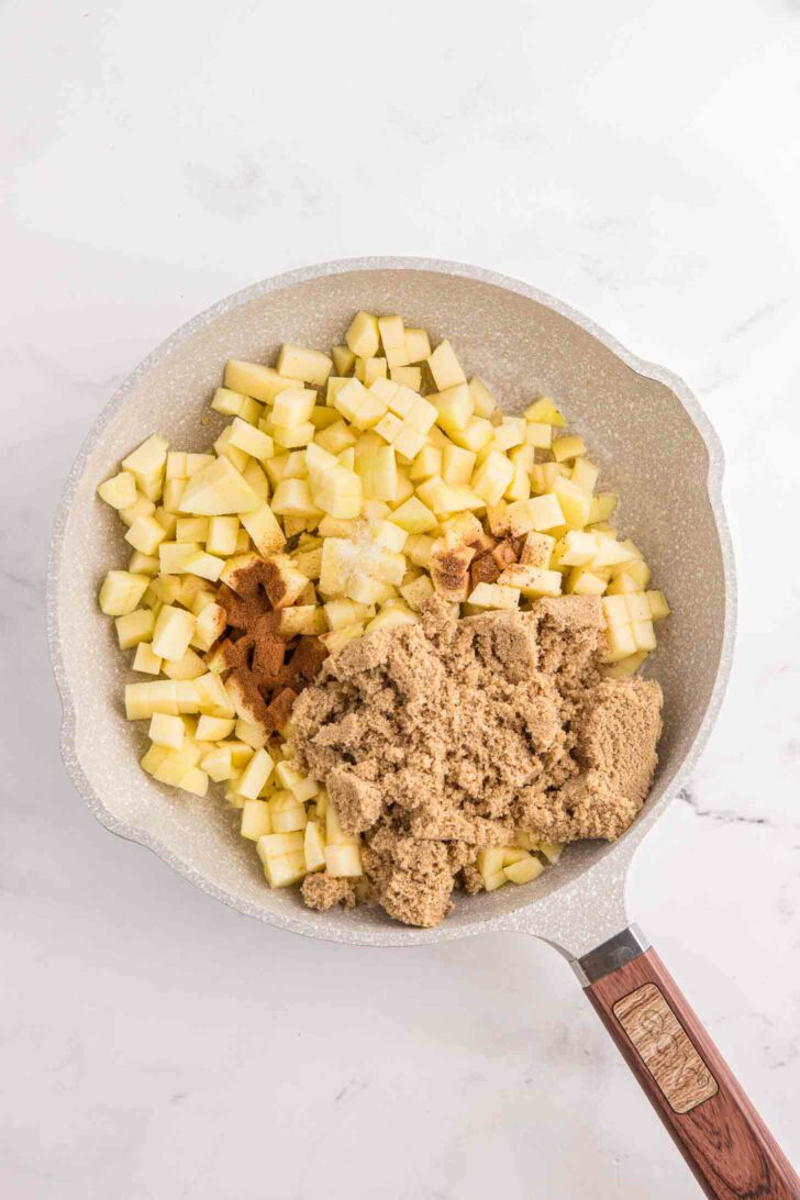 A frying pan filled with diced apples, brown sugar, and a sprinkle of cinnamon, all ready to be cooked. The pan is on a marble countertop with a wooden handle.