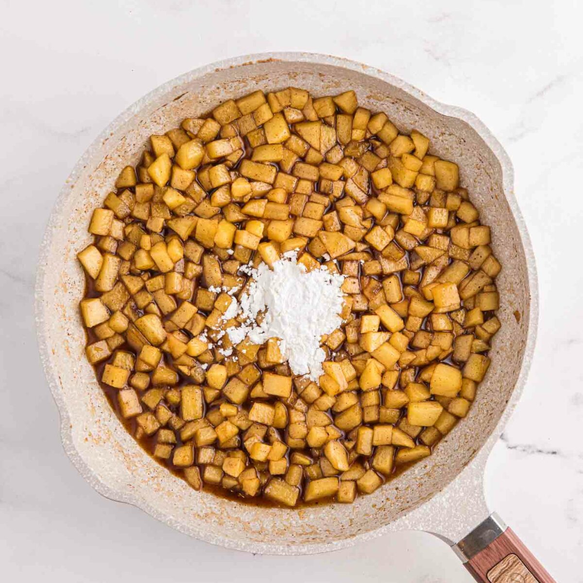A frying pan filled with diced golden-brown apples, lightly seasoned with brown sugar and spices. A small mound of cornstarch is sprinkled in the center. The pan rests on a light marble surface.