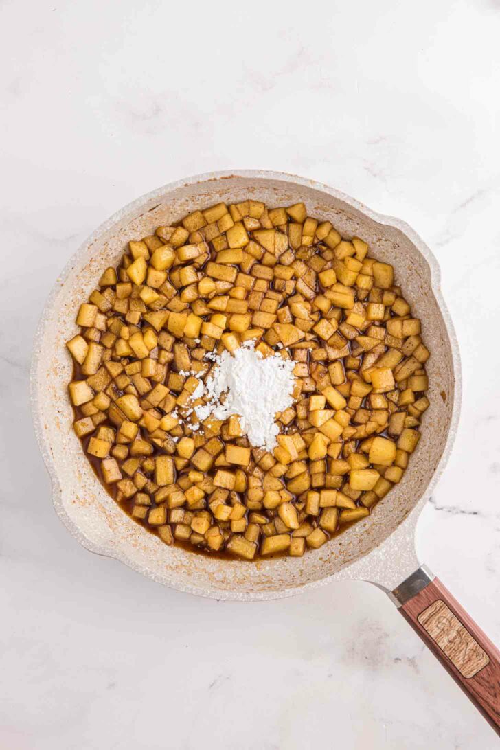 A frying pan filled with diced golden-brown apples, lightly seasoned with brown sugar and spices. A small mound of cornstarch is sprinkled in the center. The pan rests on a light marble surface.