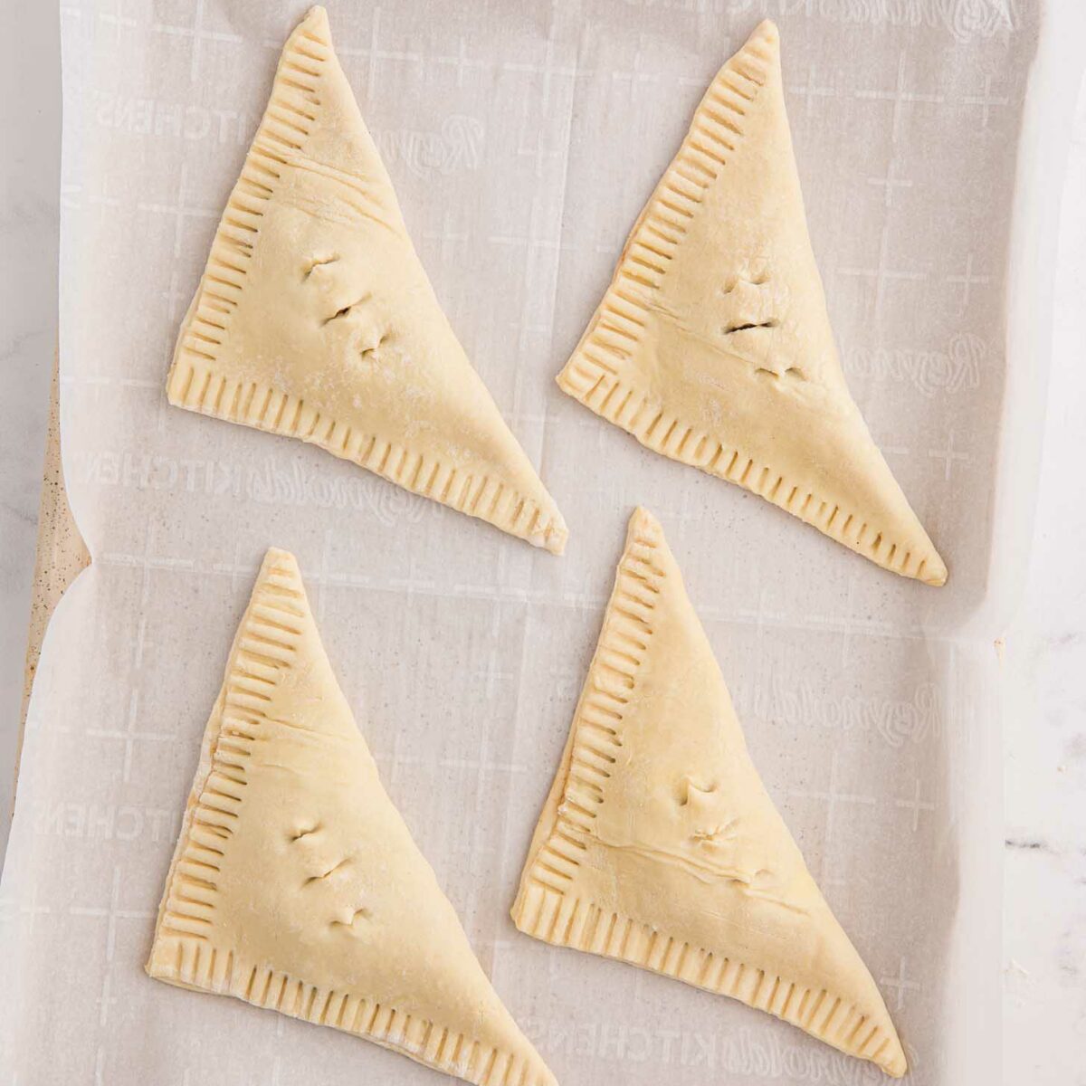 Four unbaked, triangular puff pastry turnovers lie on a parchment-lined baking sheet. Each pastry has crimped edges and small slits on top.