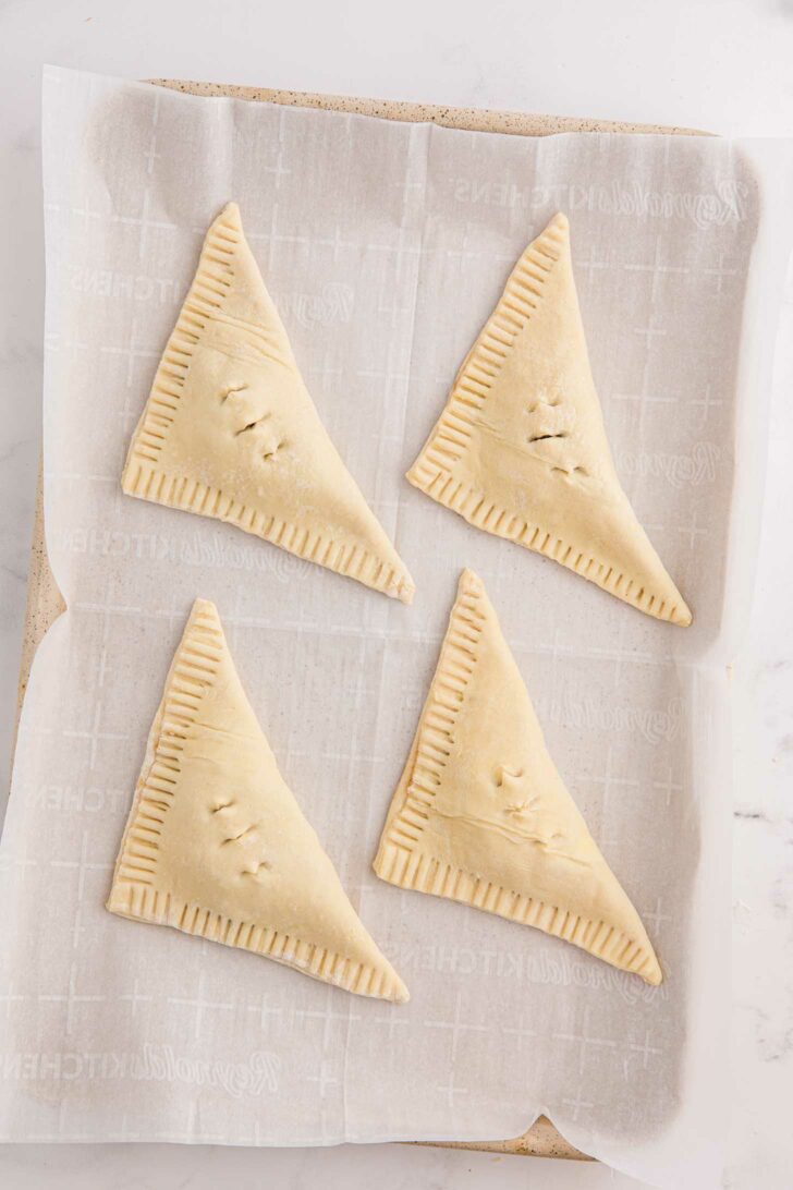 Four unbaked, triangular puff pastry turnovers lie on a parchment-lined baking sheet. Each pastry has crimped edges and small slits on top.