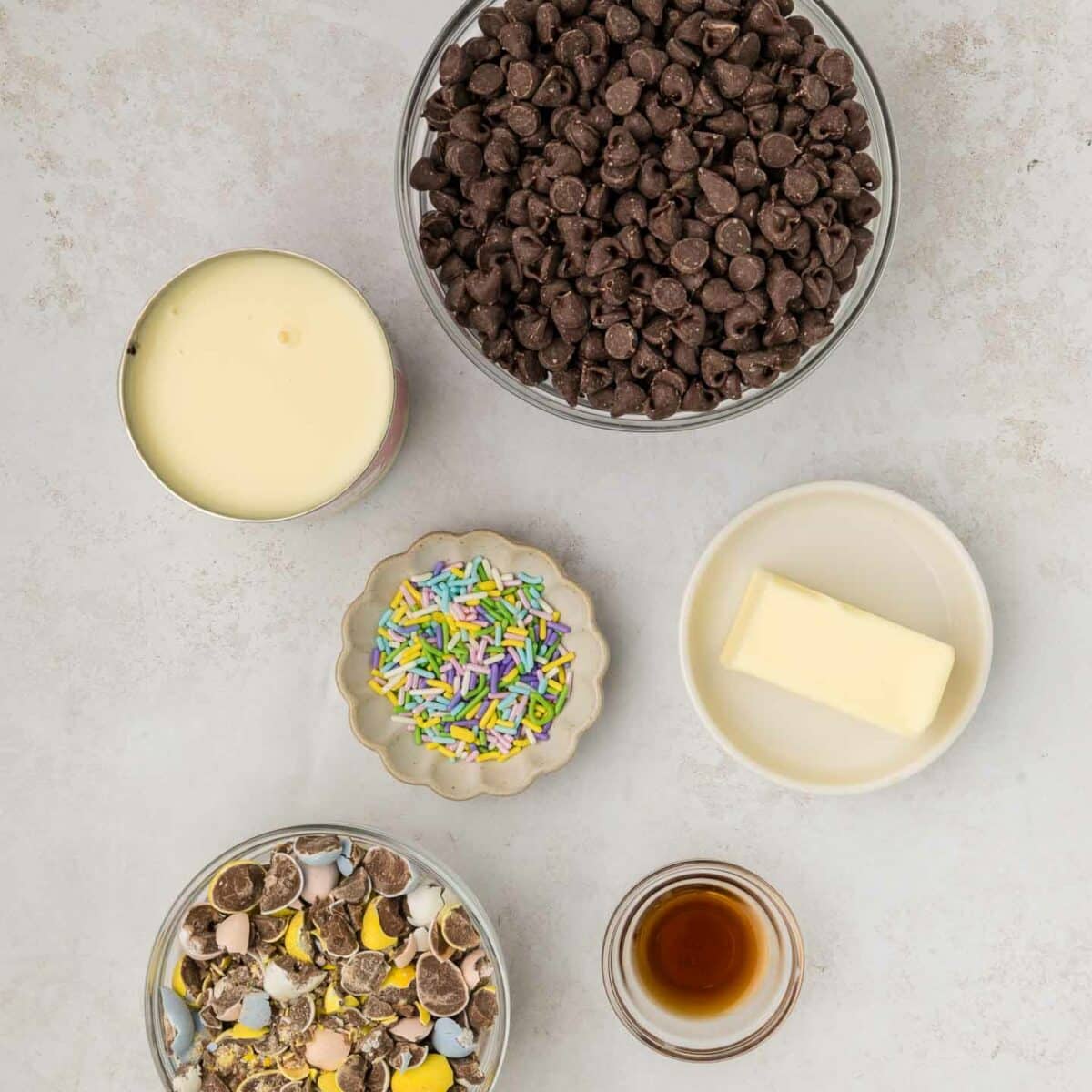 Ingredients for a recipe on a light surface include a bowl of chocolate chips, a dish of butter, a glass of vanilla extract, a bowl of colorful sprinkles, crumbled chocolate eggs, and an open can of condensed milk.