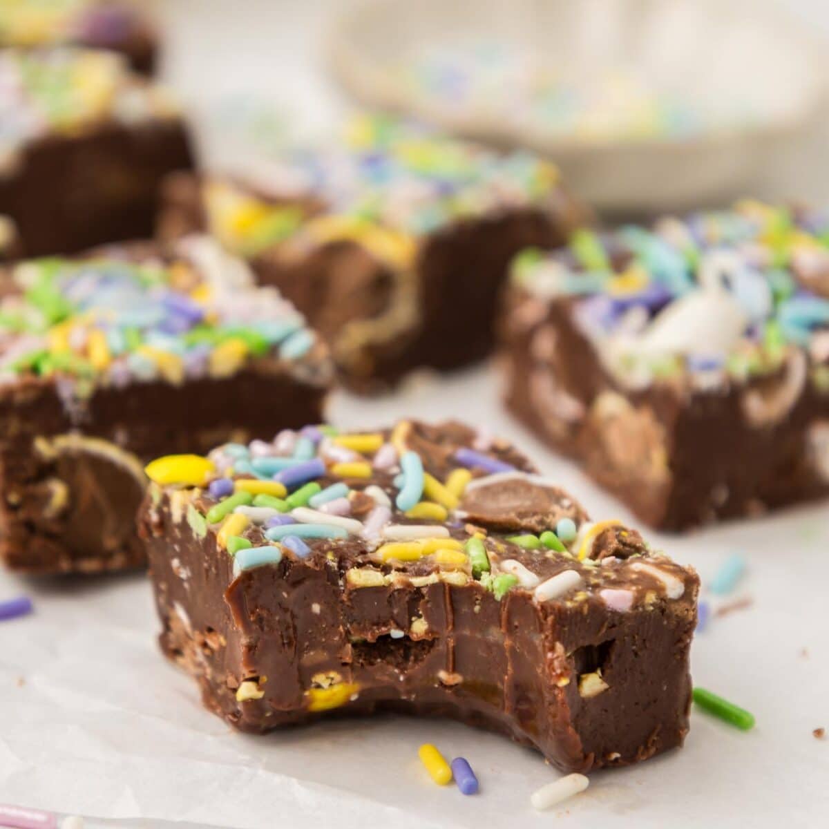 Close-up of chocolate fudge pieces with colorful sprinkles and chocolate eggs on top, arranged on white parchment paper. One piece has a bite taken out, revealing a rich, soft texture. A small dish is blurred in the background.