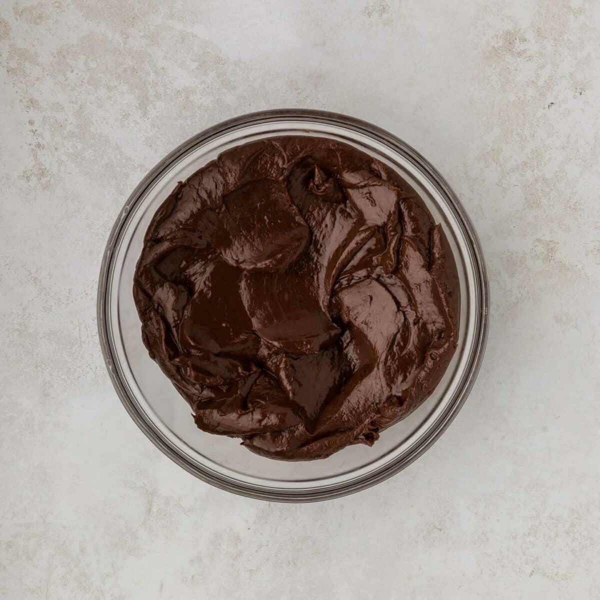 A glass bowl filled with smooth, dark chocolate frosting sits on a textured, light gray surface.
