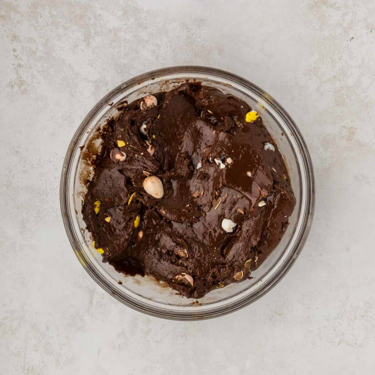 A glass bowl filled with dark chocolate mixed with chocolate eggs, placed on a light gray marble surface.