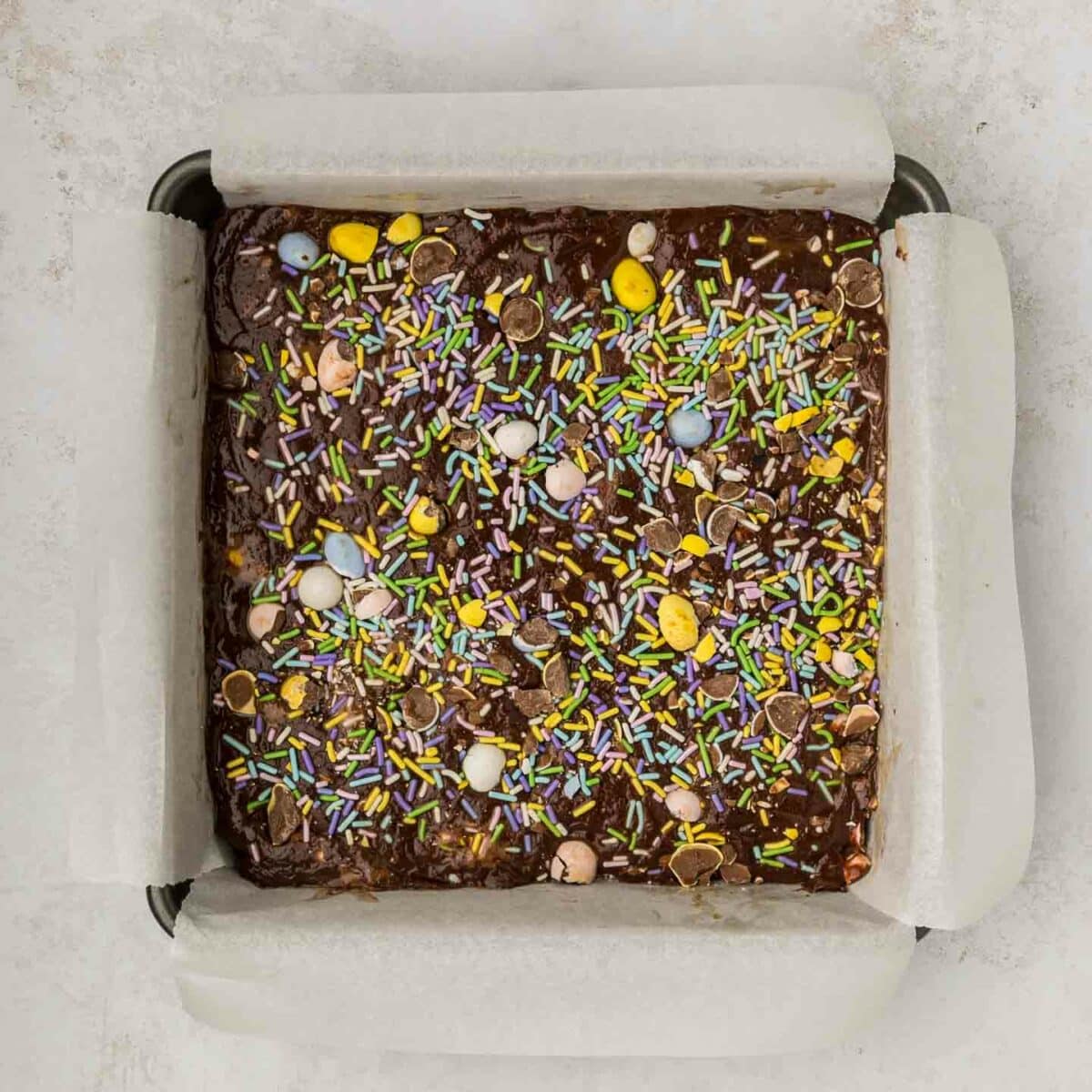 A square pan filled with chocolate fudge topped with colorful sprinkles and small pastel eggs. The pan is lined with parchment paper, set on a light-colored countertop.