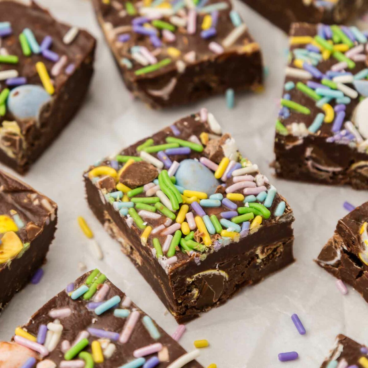 Close-up of chocolate fudge squares topped with colorful sprinkles and candy pieces, laid out on a parchment paper. The fudge has a rich, dark color with a smooth, glossy surface, and the sprinkles add a festive touch.