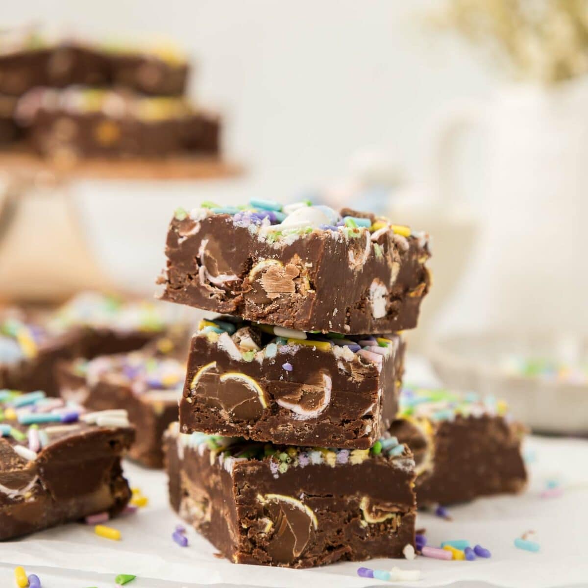 A stack of three pieces of chocolate fudge topped with colorful sprinkles and embedded with chunks of chocolate eggs. More fudge pieces are blurred in the background, with a soft focus on light tableware and a plant.