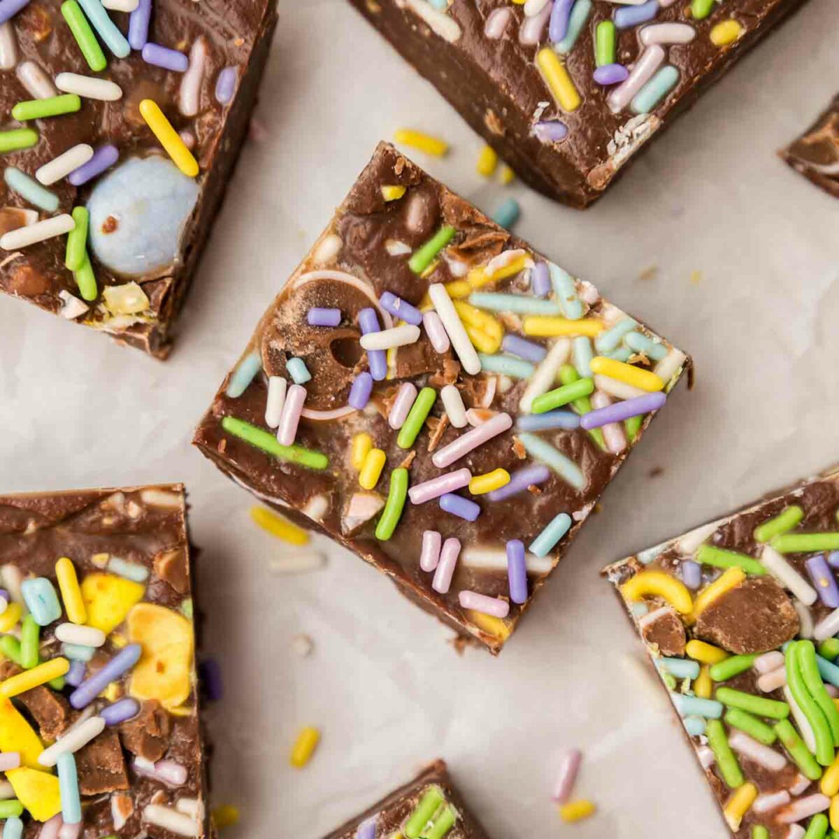 Top view of colorful chocolate squares on a white surface, sprinkled with pastel rainbow sprinkles. Each piece appears to be a rich chocolate treat, studded with additional colorful candies or cereal pieces.