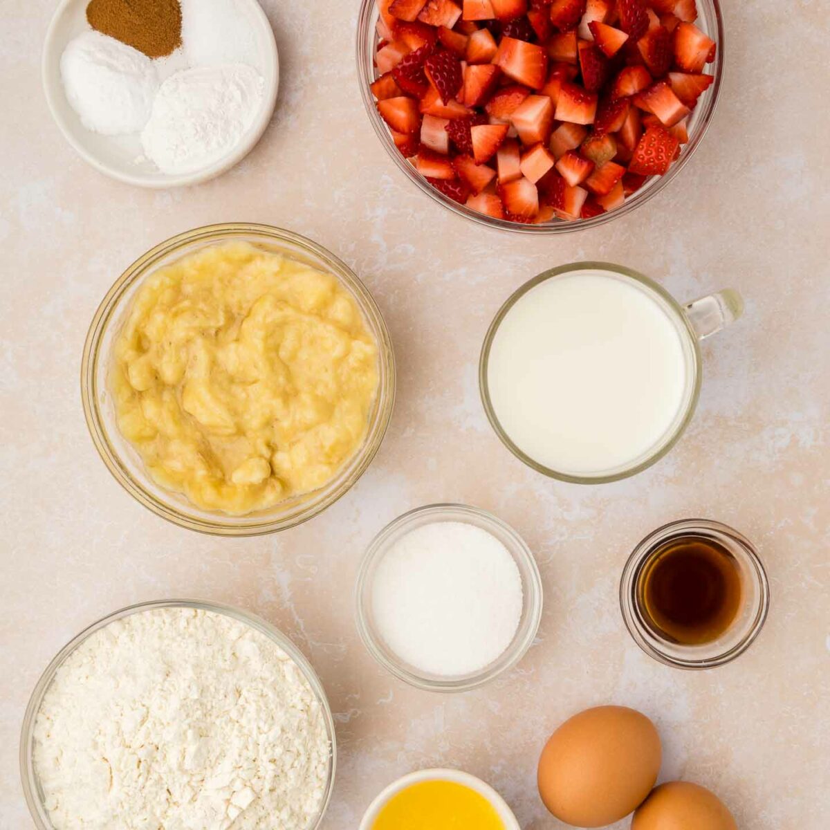Ingredients for baking are arranged on a light surface: chopped strawberries, flour, mashed bananas, milk, two eggs, melted butter, vanilla extract, sugar, and small bowls of baking powder, baking soda, and cinnamon.