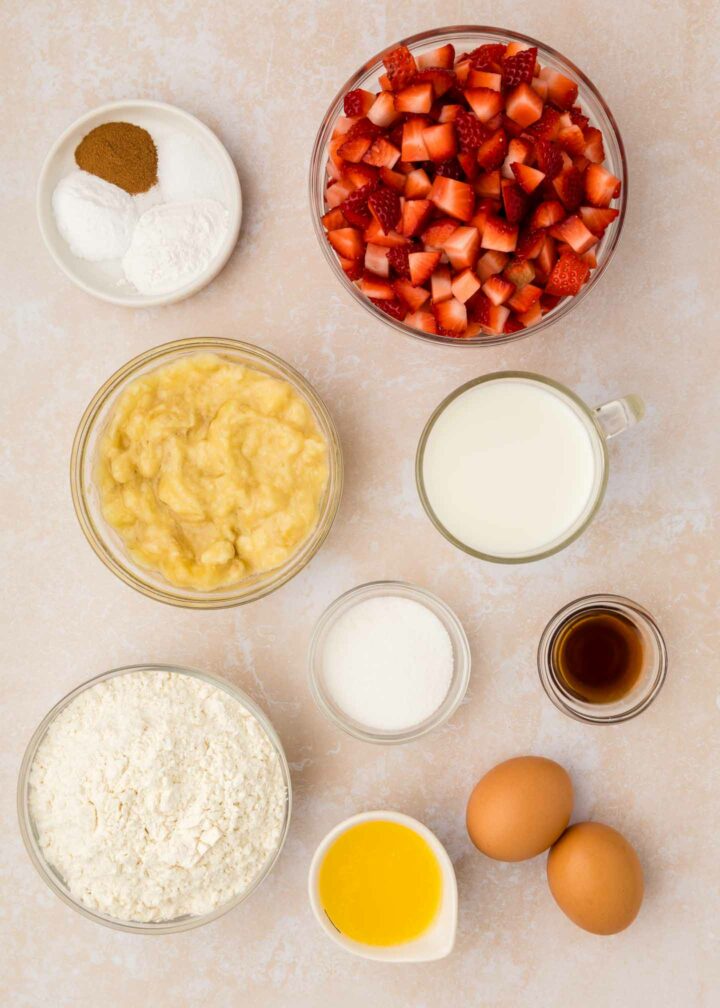 Ingredients for baking are arranged on a light surface: chopped strawberries, flour, mashed bananas, milk, two eggs, melted butter, vanilla extract, sugar, and small bowls of baking powder, baking soda, and cinnamon.
