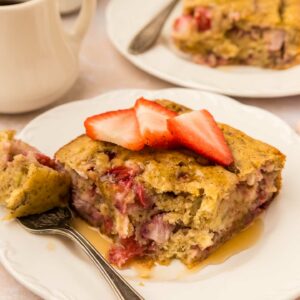 Two slices of strawberry banana pancake casserole on white plates, topped with sliced strawberries and drizzled with syrup. A fork is on one plate, and a cup of coffee is in the background.