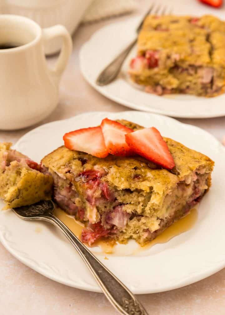 Two slices of strawberry banana pancake casserole on white plates, topped with sliced strawberries and drizzled with syrup. A fork is on one plate, and a cup of coffee is in the background.
