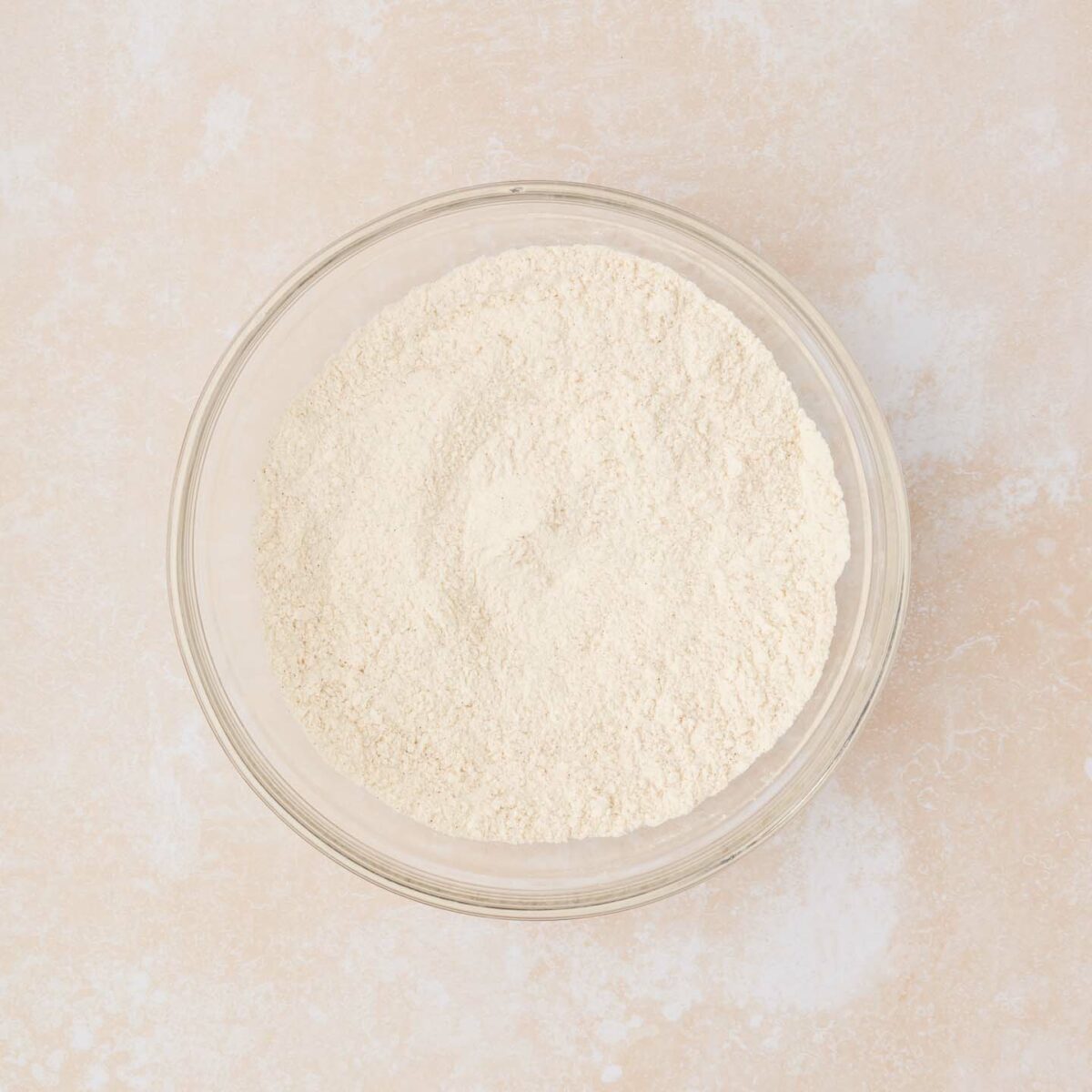 A clear glass bowl filled with a flour mixture sits on a textured beige surface.