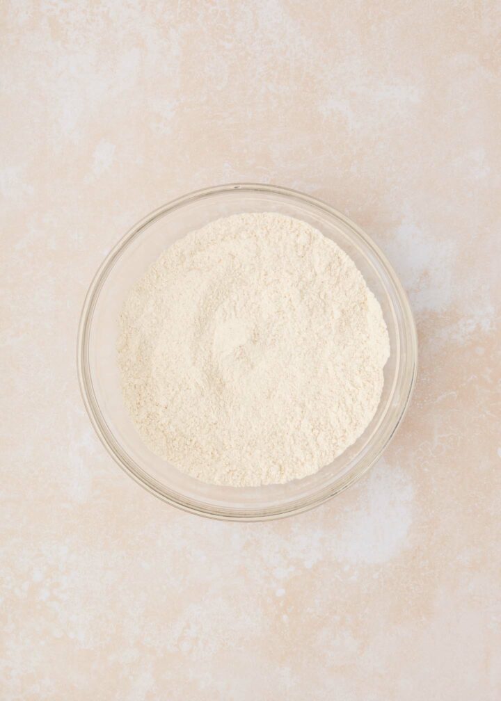 A clear glass bowl filled with a flour mixture sits on a textured beige surface.