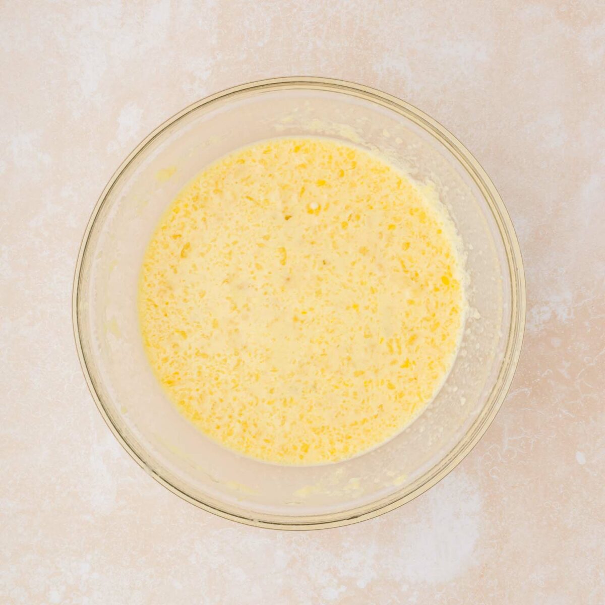 A clear glass bowl filled with a mixture of beaten eggs and wet ingredients, visible with bubbles and light froth on top, sits on a beige countertop.