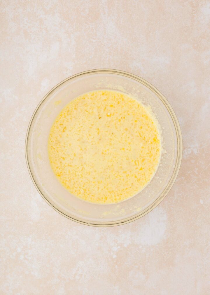 A clear glass bowl filled with a mixture of beaten eggs and wet ingredients, visible with bubbles and light froth on top, sits on a beige countertop.