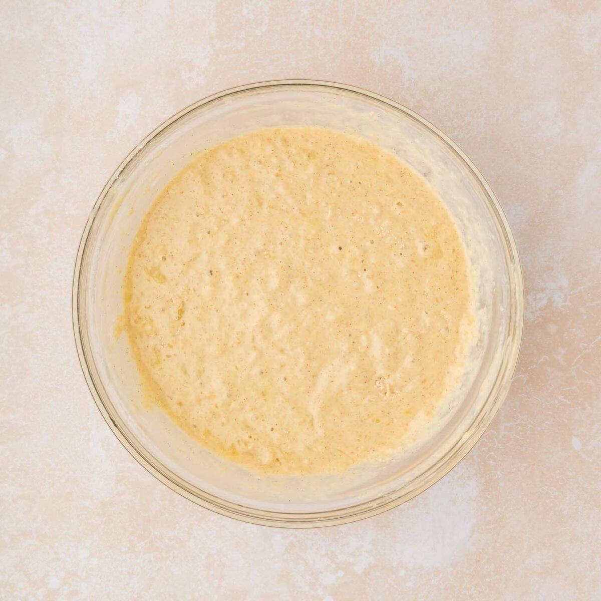 A clear glass bowl filled with light yellow, frothy batter sits on a textured, light beige countertop. The batter appears bubbly and smooth.