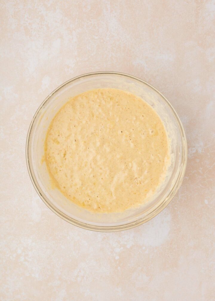 A clear glass bowl filled with light yellow, frothy batter sits on a textured, light beige countertop. The batter appears bubbly and smooth.