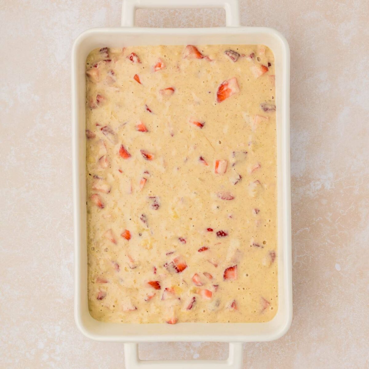 A rectangular baking dish filled with a thick, creamy pancake batter mixed with chopped red strawberries. The dish rests on a light beige textured surface.