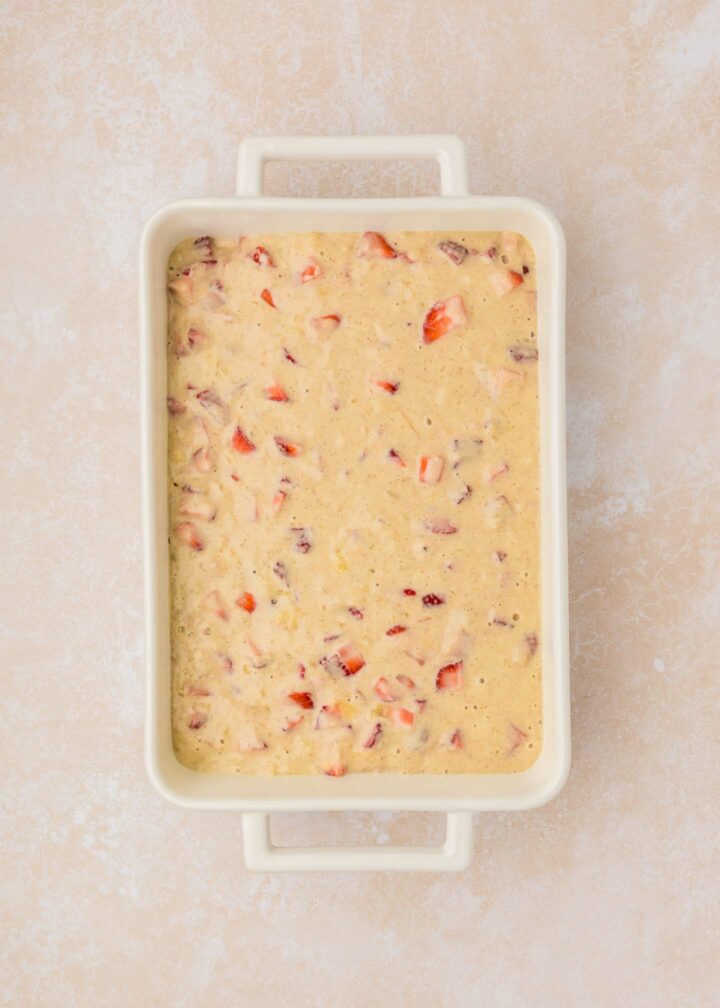 A rectangular baking dish filled with a thick, creamy pancake batter mixed with chopped red strawberries. The dish rests on a light beige textured surface.