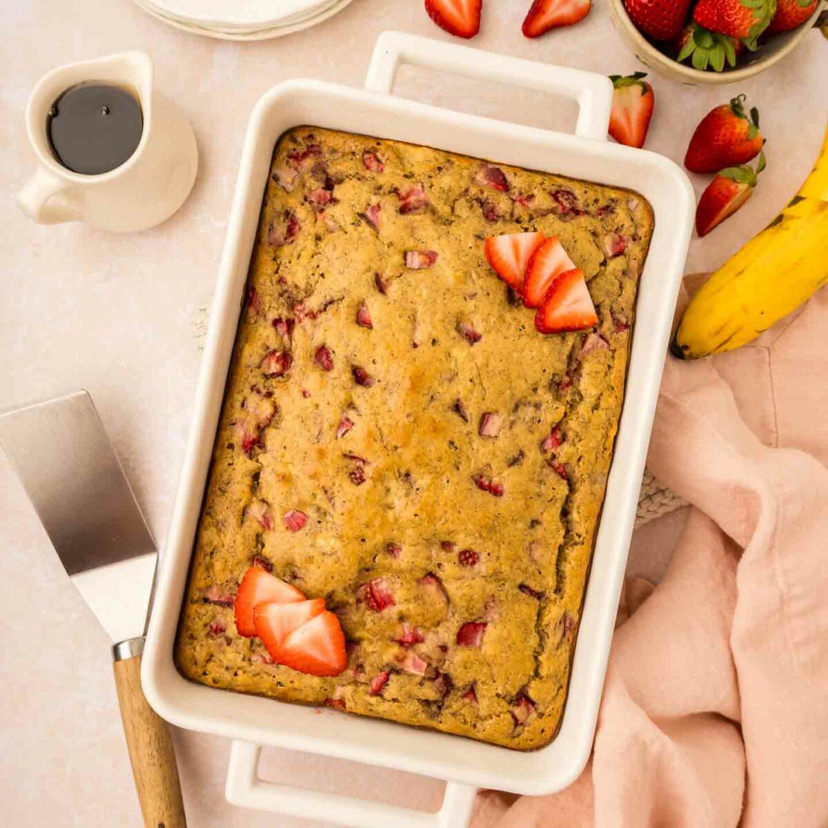 A rectangular strawberry banana pancake casserole in a white baking dish is on a table. Sliced strawberries garnish the top. Side items include a pink cloth, a cake server, a banana, a cup of coffee, and a bowl of fresh strawberries.