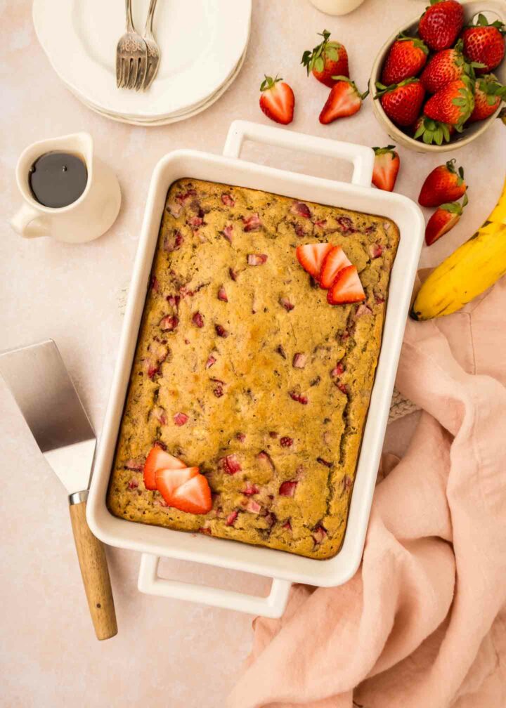 A rectangular strawberry banana pancake casserole in a white baking dish is on a table. Sliced strawberries garnish the top. Side items include a pink cloth, a cake server, a banana, a cup of coffee, and a bowl of fresh strawberries.