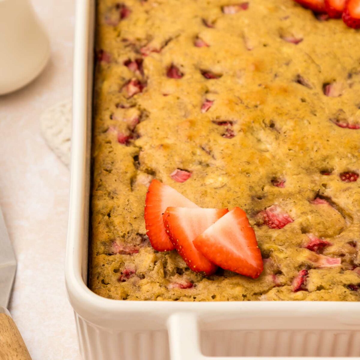 Close-up of a rectangular strawberry banana pancake casserole in a white dish. The casserole is golden brown with pieces of strawberries visible. Sliced strawberries garnish the top. A knife and cup are seen in the blurred background.