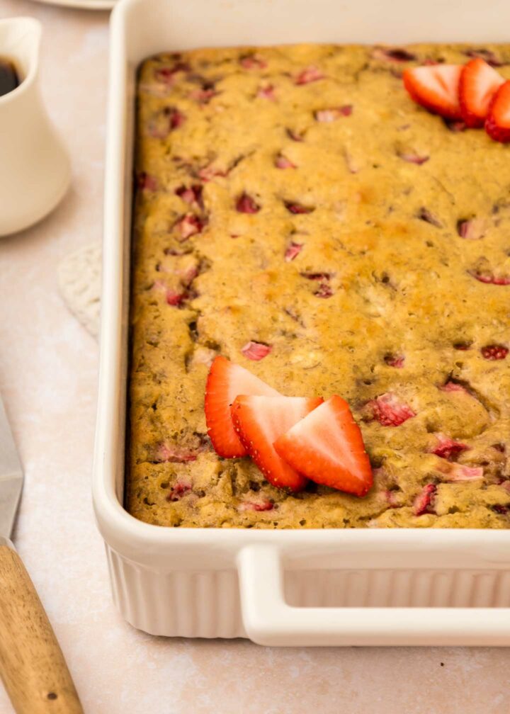 Close-up of a rectangular strawberry banana pancake casserole in a white dish. The casserole is golden brown with pieces of strawberries visible. Sliced strawberries garnish the top. A knife and cup are seen in the blurred background.