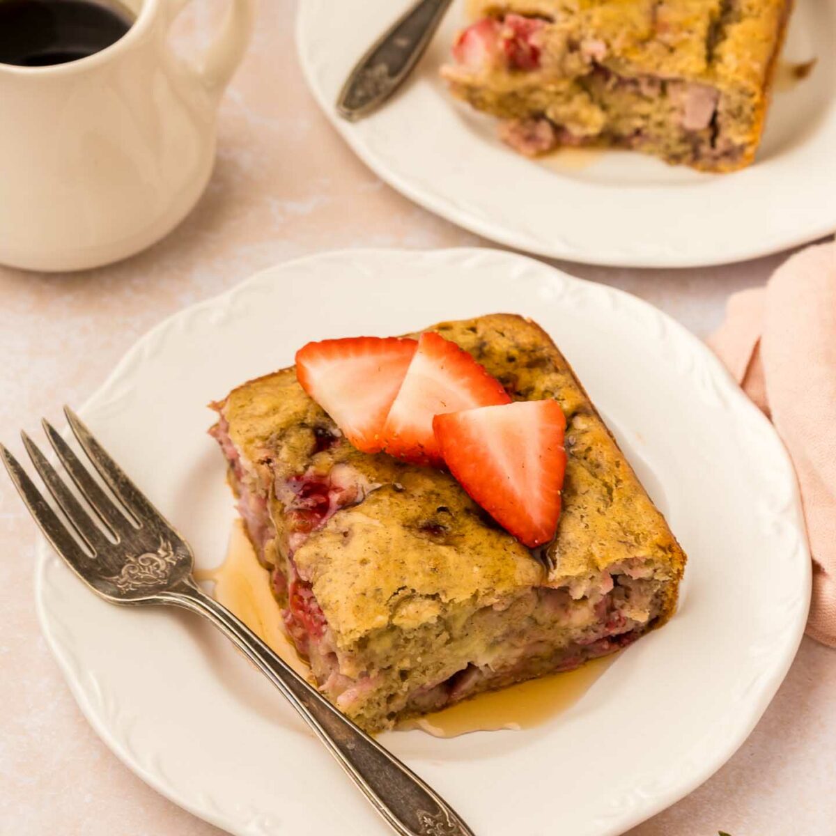 A slice of strawberry banana pancake casserole is served on a white plate, topped with fresh strawberry slices. A fork rests beside it. In the background, theres a cup of black coffee and another plate with a similar slice.