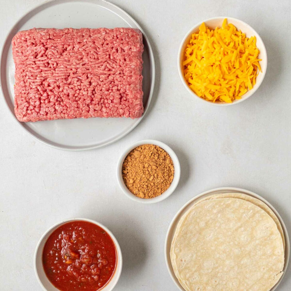 Raw ground meat on a plate, shredded cheddar cheese in a bowl, taco seasoning, salsa, and corn tortillas laid out on a light surface.