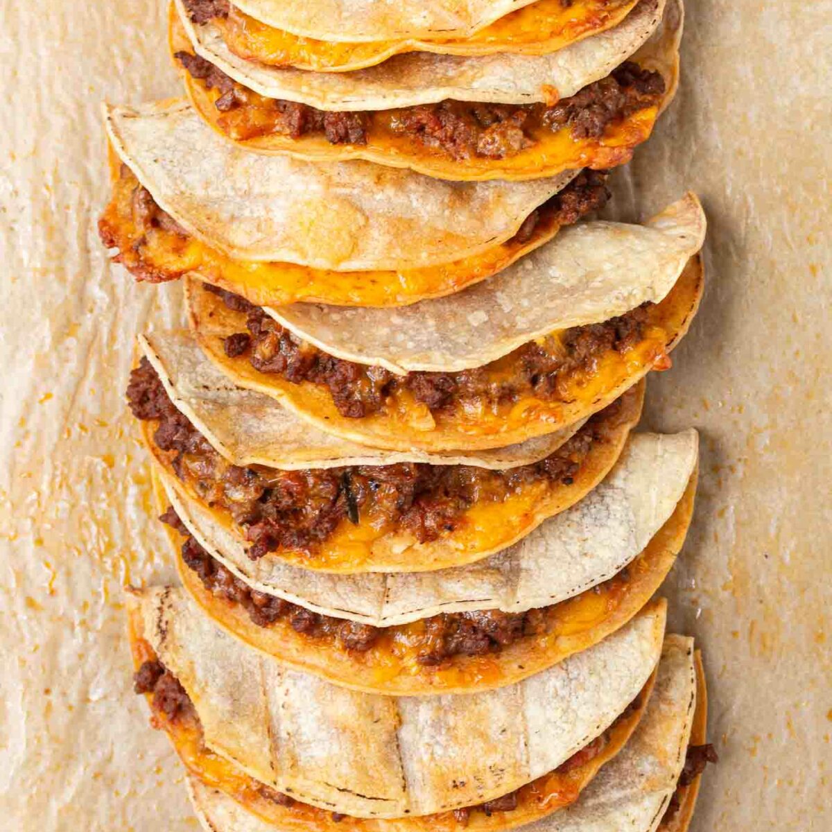 A stack of crispy tacos filled with seasoned ground meat and melted cheese. The tacos are arranged in a row on a parchment-covered surface, with bowls of sliced tomatoes and olives partially visible at the corners.