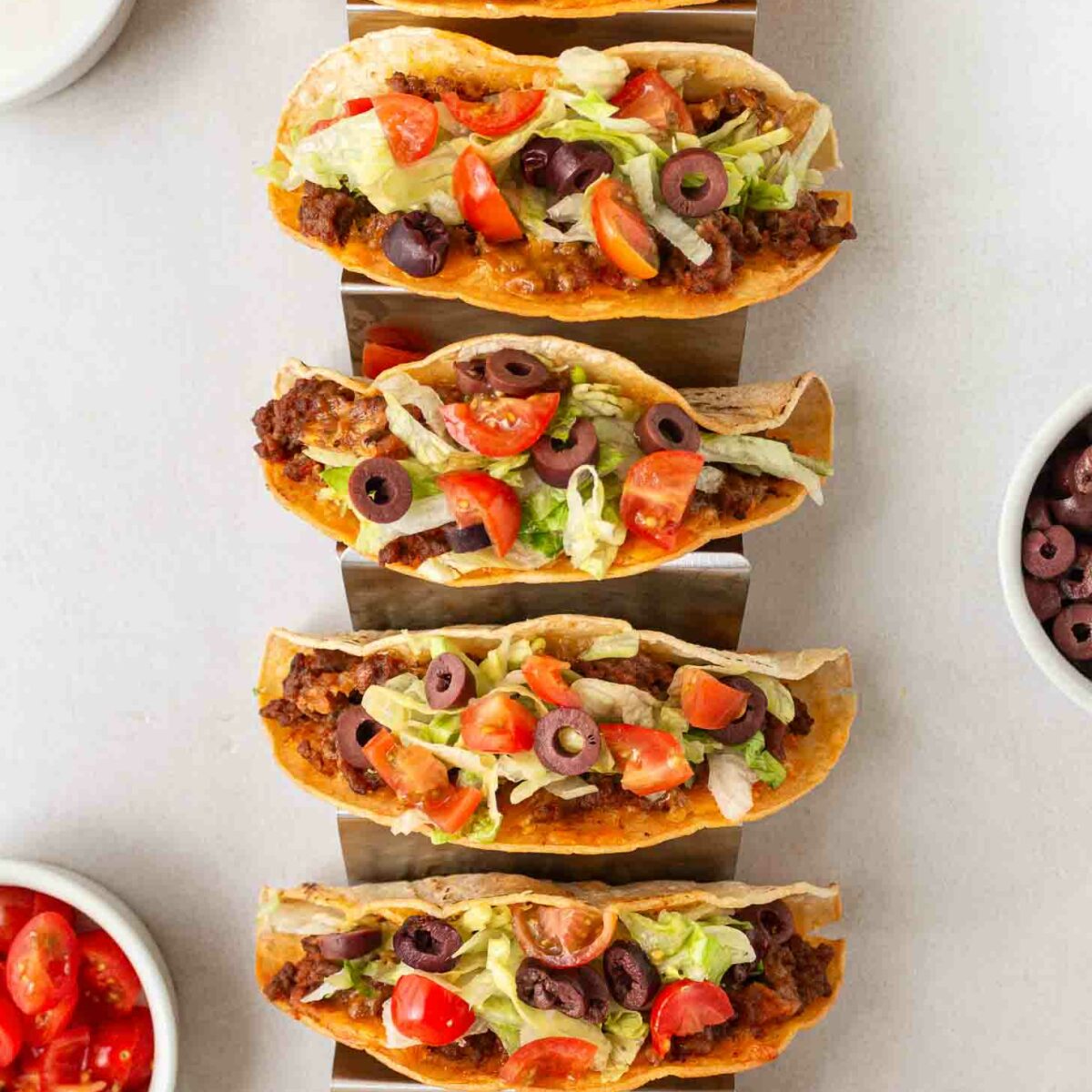 Six beef tacos with melted cheese, lettuce, tomatoes, and olives are lined up in metal holders. Bowls of chopped tomatoes and black olives are at the side. The scene is set on a light gray surface.