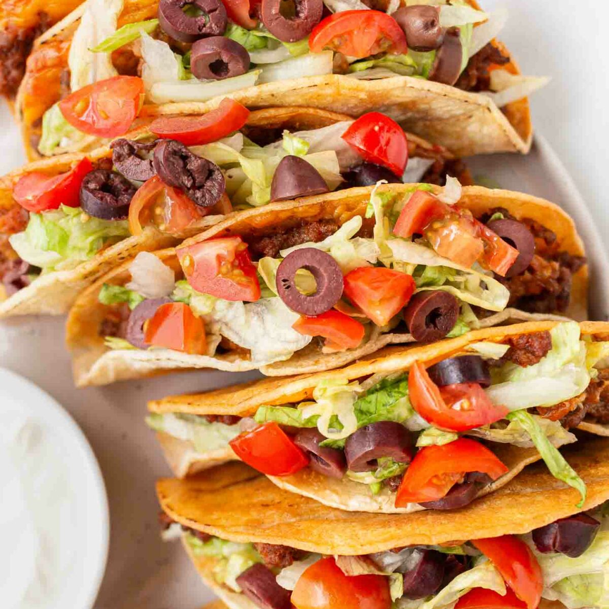 A plate of crispy tacos filled with seasoned ground beef, shredded lettuce, diced tomatoes, sliced black olives, and melted cheese. A small bowl of sliced black olives is partially visible in the background.