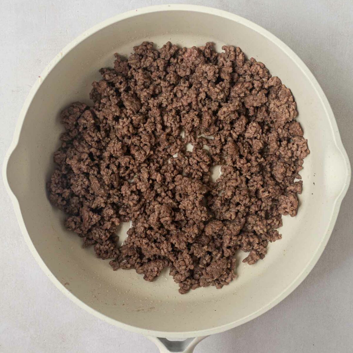 A white frying pan with a wooden handle contains browned ground beef, evenly spread across the surface, on a light gray countertop.