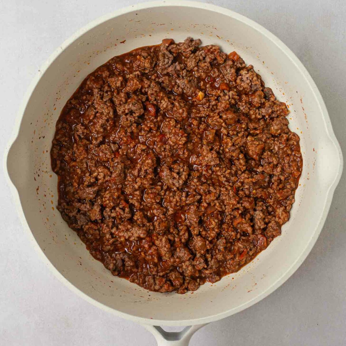 A white ceramic pan filled with cooked ground beef with taco seasoning and salsa. The meat is finely crumbled and evenly distributed across the pan surface. The pan has a wooden handle and is set on a light gray background.