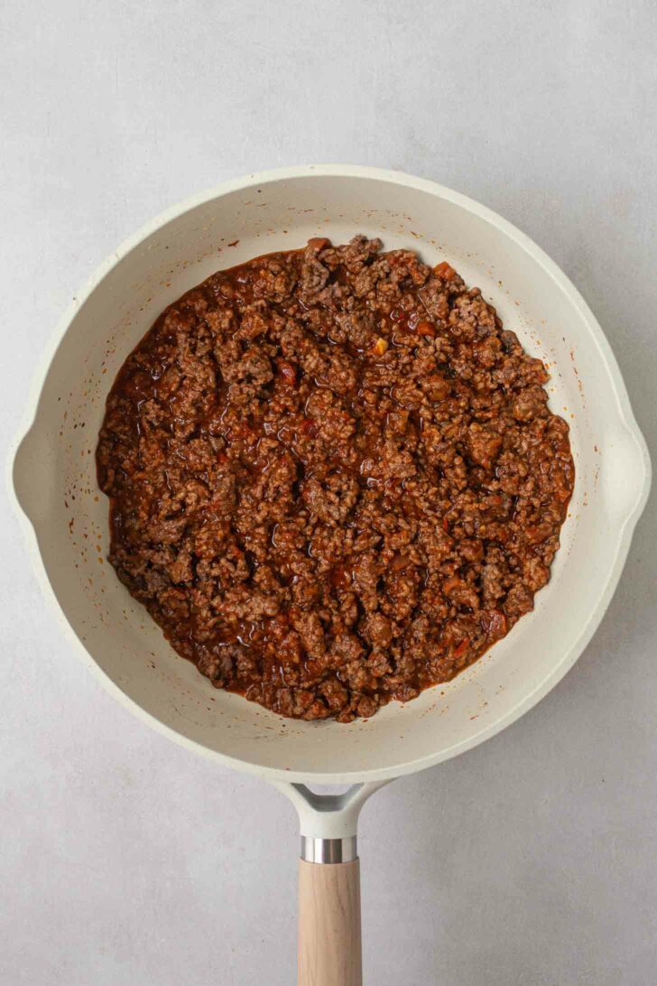 A white ceramic pan filled with cooked ground beef with taco seasoning and salsa. The meat is finely crumbled and evenly distributed across the pan surface. The pan has a wooden handle and is set on a light gray background.