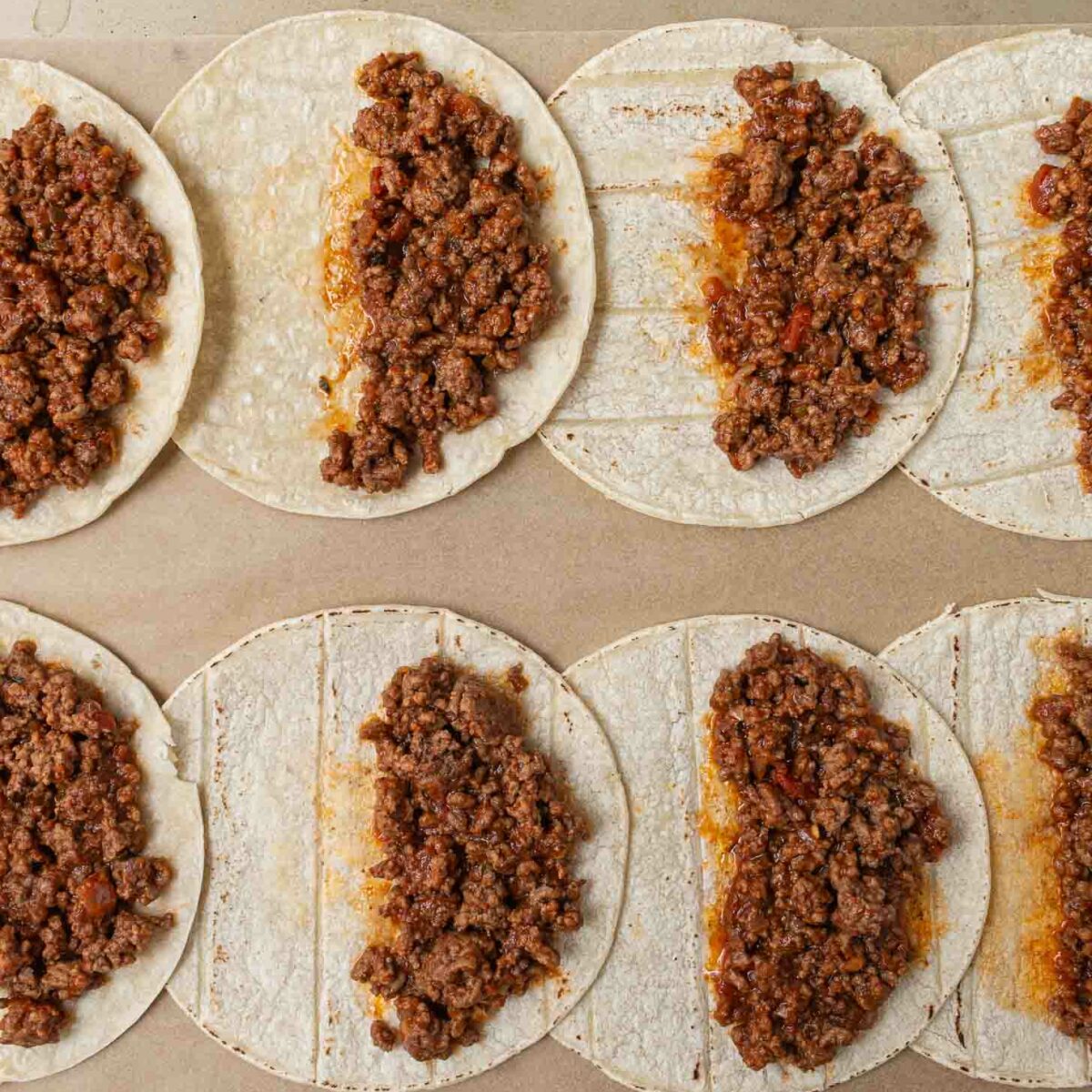 A baking tray with eight corn tortillas, each topped with seasoned ground beef filling on one half, ready to be folded or baked. The tray is on a neutral background.
