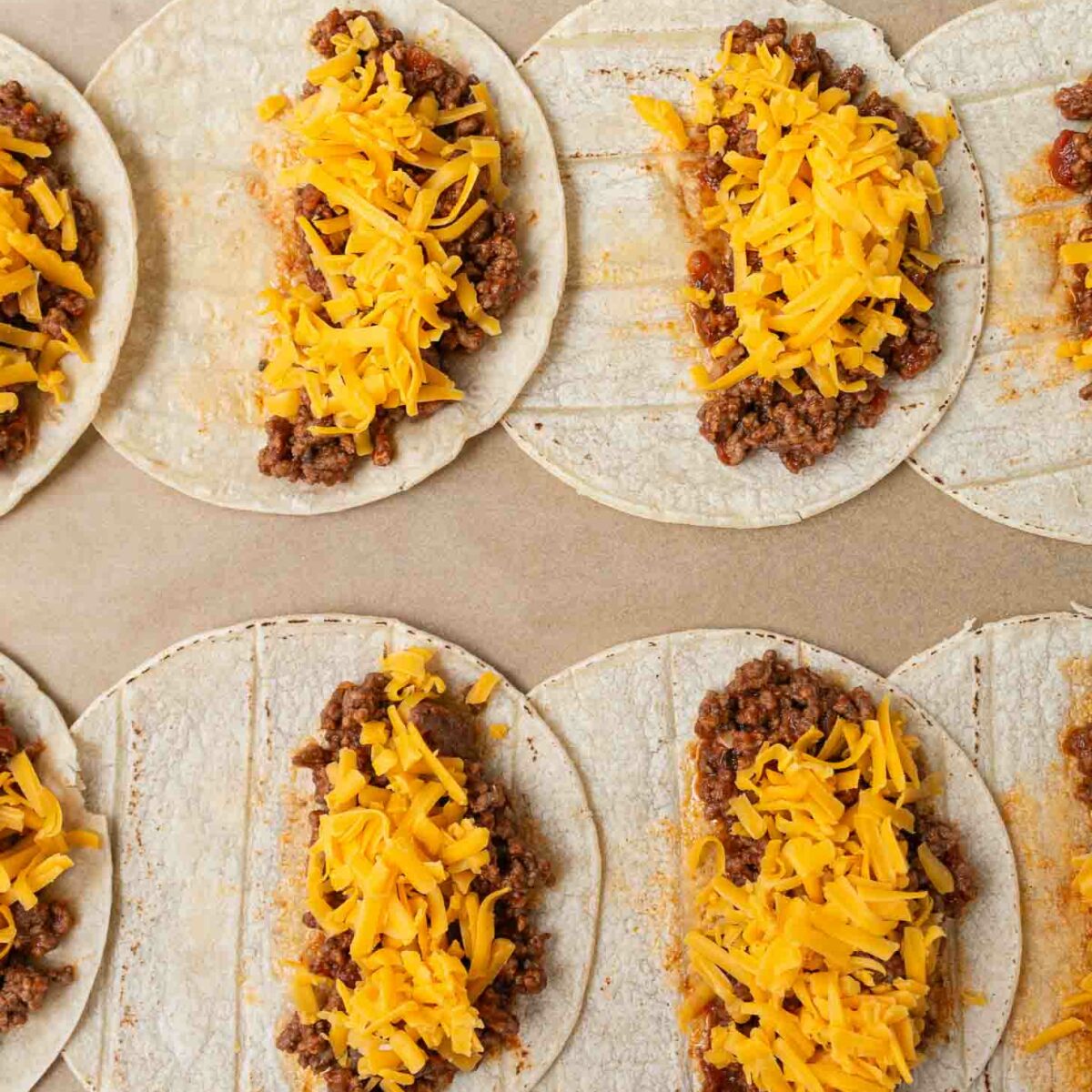 Corn tortillas lined up on a baking sheet, each filled with seasoned ground beef and topped with shredded cheddar cheese. The fillings are arranged in a line down the center of each tortilla.